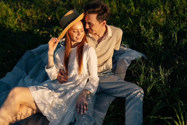 Romantica giovane coppia sorridente innamorata con gli occhi chiusi seduti abbracciati insieme sul bellissimo prato verde nella sera d'estate durante il tramonto dorato