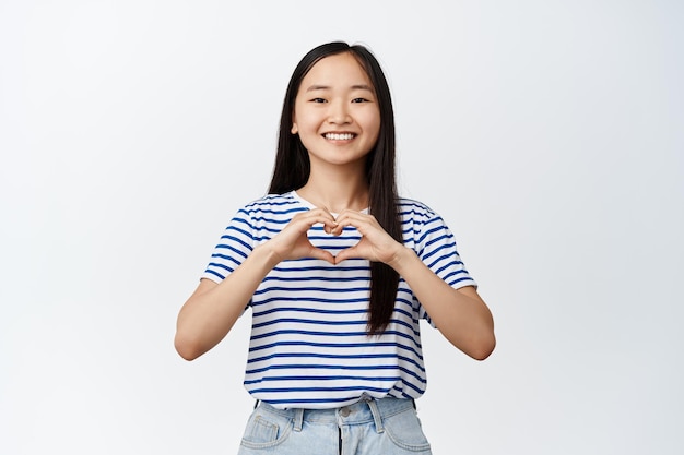 Romantica ragazza asiatica mostra il segno del cuore e sorrisi happe alla fotocamera in piedi su sfondo bianco Copia spazio