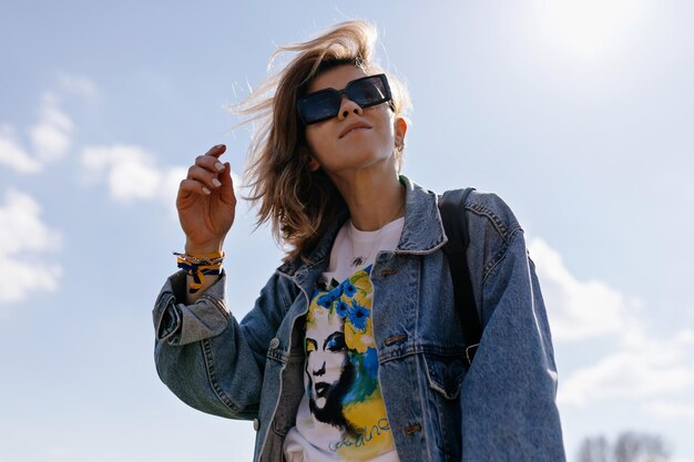 Romantica ragazza dai capelli biondi sta camminando in una giornata di sole e distoglie lo sguardo contro il cielo blu Closeup ritratto di spensierata giovane donna in giacca di jeans sta camminando fuori