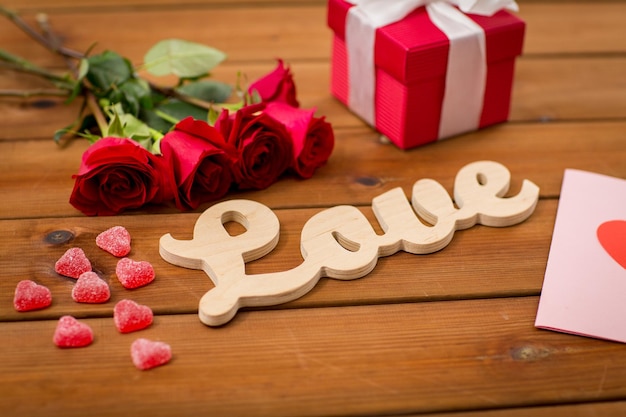 romanticismo, concetto di San Valentino e vacanze - primo piano della parola amore, confezione regalo, rose rosse e biglietto di auguri con caramelle a forma di cuore su legno