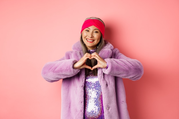 Romanticismo e San Valentino. Felice donna anziana asiatica che mostra il segno del cuore, ti amo gesto, sorridente e guardando la telecamera premurosa, in piedi in pelliccia sintetica, sfondo rosa.