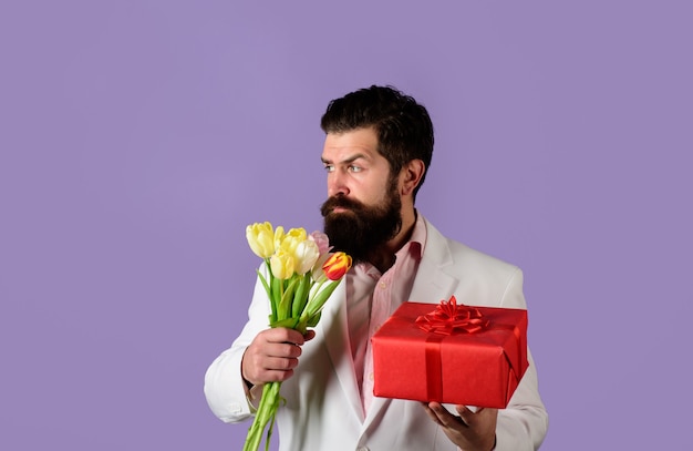 Romantico uomo barbuto tiene un mazzo di fiori e una confezione regalo il giorno di san valentino il compleanno delle donne