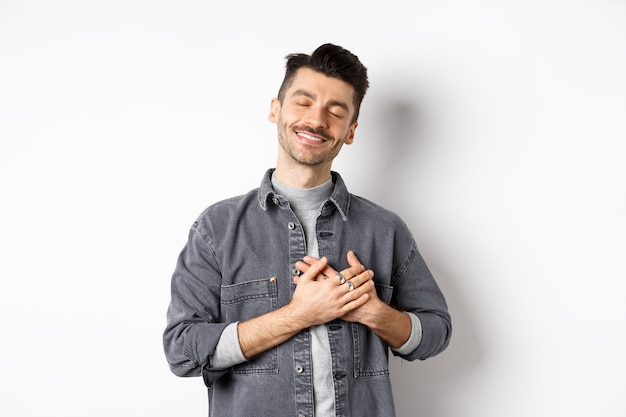 Romantico uomo felice mano nella mano sul cuore e sorridente con gli occhi chiusi, sognando qualcosa, sentendosi nostalgico da dolci ricordi, in piedi su sfondo bianco.