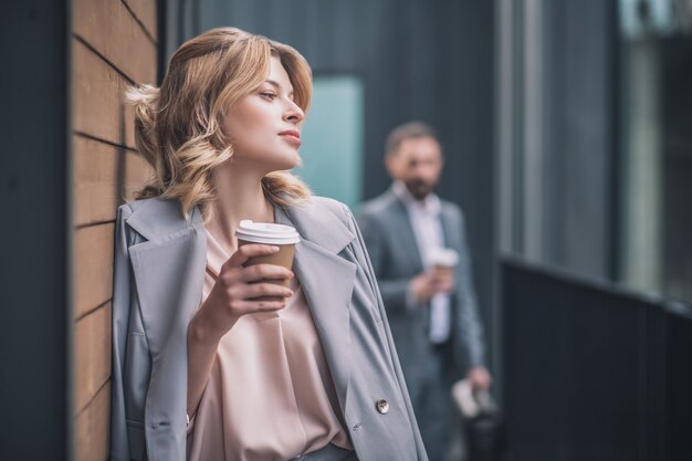 Rompere. Bella giovane donna bionda in giacca di affari in piedi pensieroso con caffè appoggiato contro il muro di edificio per uffici e uomo