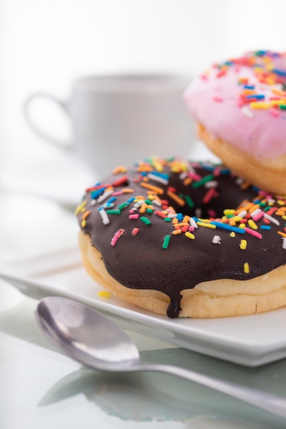 Rosa e cioccolato spruzza ciambelle su un piatto bianco e una tazza di caffè su una luce.