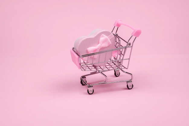 Rosa grande regalo a forma di cuore con fiocco per San Valentino nello shopping scart su uno sfondo rosa, minimalismo.