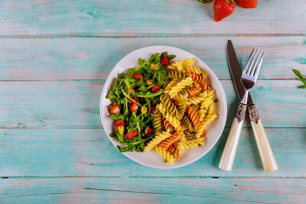 Rotini di pasta con insalata di rucola e fragole con forchetta e coltello su superficie di legno Vista dall'alto