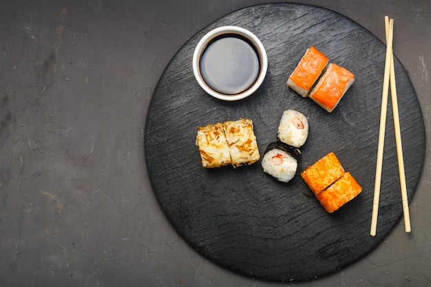 Rotoli assortiti su tavola nera accanto a bastoncini, salsa di soia e zenzero su fondo scuro.