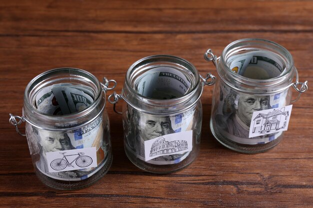 Rotoli di banconote in dollari per esigenze diverse in vasetti di vetro su sfondo di legno