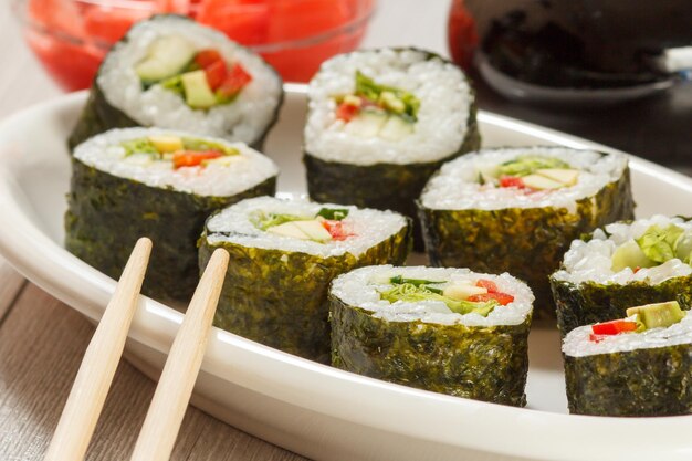 Rotoli di sushi con riso, pezzi di avocado, cetriolo, peperone rosso e foglie di lattuga su piatto di ceramica, bacchette, bottiglia di vetro con salsa di soia e zenzero sottaceto sullo sfondo. Cibo vegetariano.