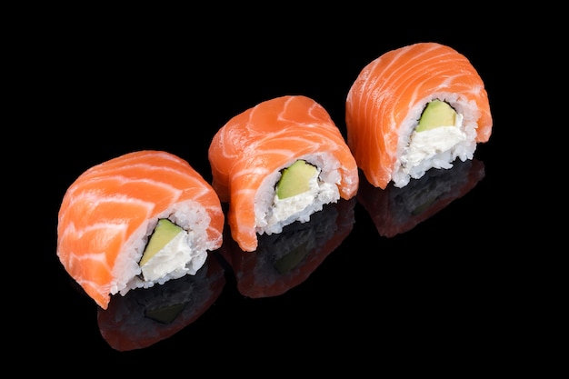 Rotoli di sushi fatti di salmone crudo fresco, crema di formaggio e avocado isolato su fondo nero con riflessi. Philadelphia, sushi tradizionale con salmone, avocado e formaggio. Cucina giapponese.