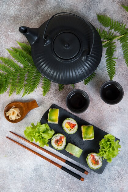 Rotoli di sushi vegetariano vegetale con una teiera di tè e tazze cinesi stanno su un tavolo di cemento. Orientamento verticale