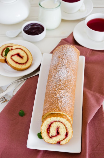 rotolo di pan di spagna con marmellata di lamponi su un piatto bianco con una tazza di tè ai frutti di bosco profumato