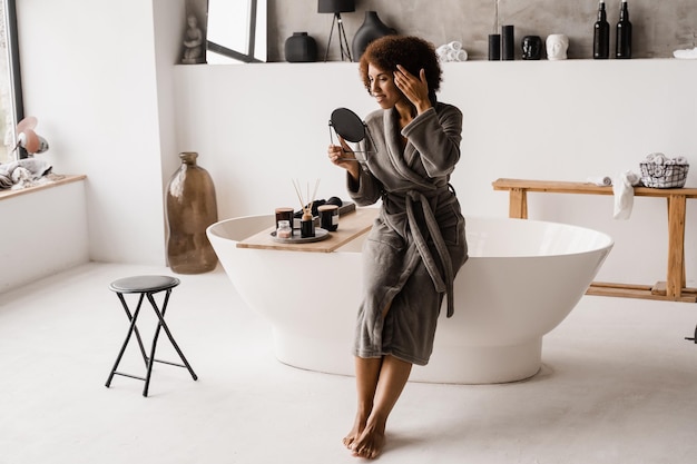 Routine mattutina di bellezza della ragazza africana in bagno Attraente donna afroamericana in accappatoio si guarda allo specchio