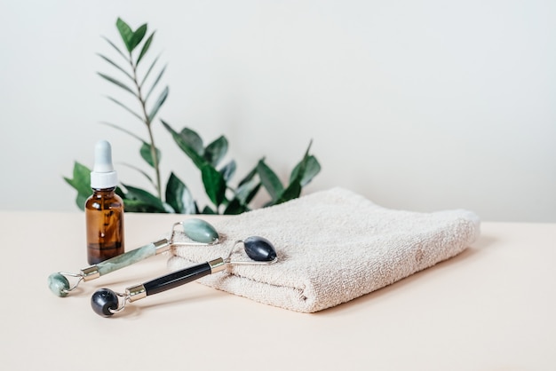 Rulli per massaggi Jade guasha sull'asciugamano e bottiglia di siero su sfondo pastello chiaro, relax SPA, concetto di cura del viso e della pelle