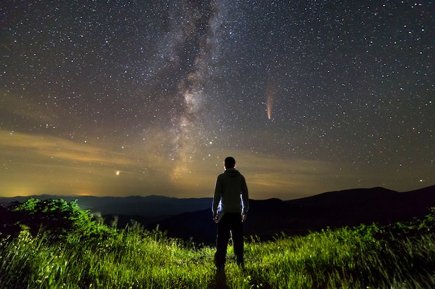 Sagoma scura di un uomo in piedi in montagna di notte godendo la Via Lattea e la cometa Neowise con coda leggera nella vista del cielo scuro.