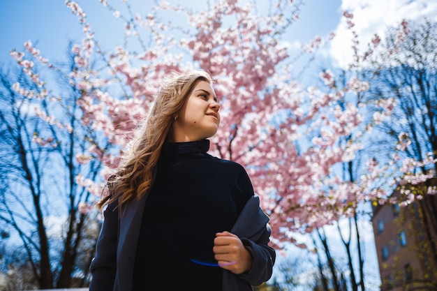 Sakura rami con fiori su un albero per le strade della città. La ragazza alla moda della donna sta nella via con la fioritura di sakura. Ragazza alla moda moderna all'aperto. Fiori dell'albero di Sakura.