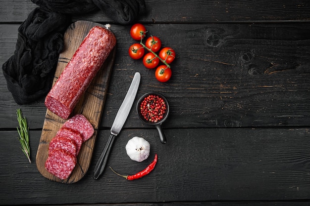 Salame secco con rosmarino fresco e set di spezie, su tavolo di legno nero, vista dall'alto piatta