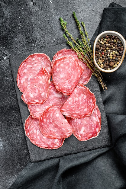 Salchichon spagnolo del salame su un tagliere nero. Sfondo nero. Vista dall'alto