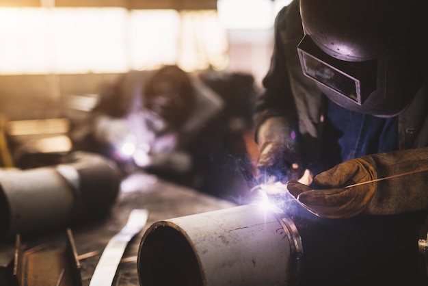 Saldatore in uniforme protettiva e maschera saldatura tubo metallico sul tavolo industriale mentre volano scintille.