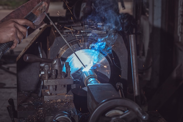 Saldatore professionale del primo piano in uniforme protettiva di sicurezza e tubo metallico del metallo di saldatura della maschera sulla tavola industriale dietro nella fabbrica industriale.