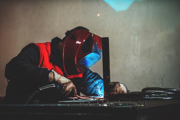 Saldatore professionale in uniforme protettiva e maschera per saldatura tubo metallico sul tavolo industriale con altri lavoratori dietro nell'officina industriale