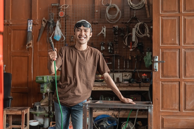 Saldatore sorridente esamina la macchina fotografica mentre tiene un saldatore elettrico in un negozio di saldatura