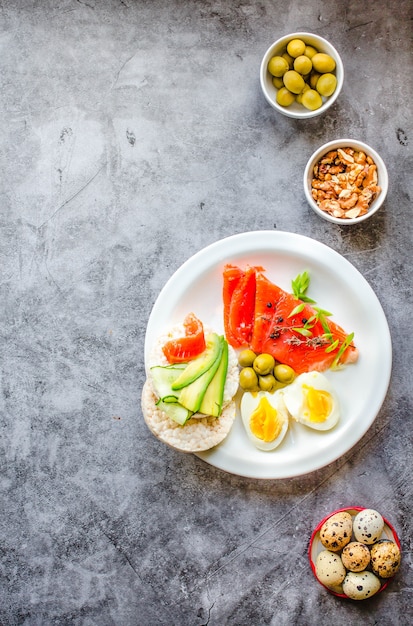 Salmone affumicato di pesce rosso con olive, uova di quaglia, noci e torte croccanti di riso con avocado su sfondo grigio. Vista dall'alto. Rustico. Prodotti con omega-3 buoni per capelli e pelle sani. Spazio per il testo.