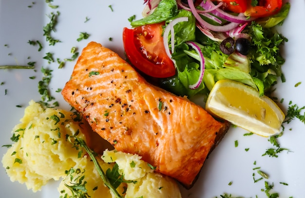 Salmone al forno con purè di patate, limone, pomodori, cipolle rosse, olive e foglie di insalata su sfondo bianco