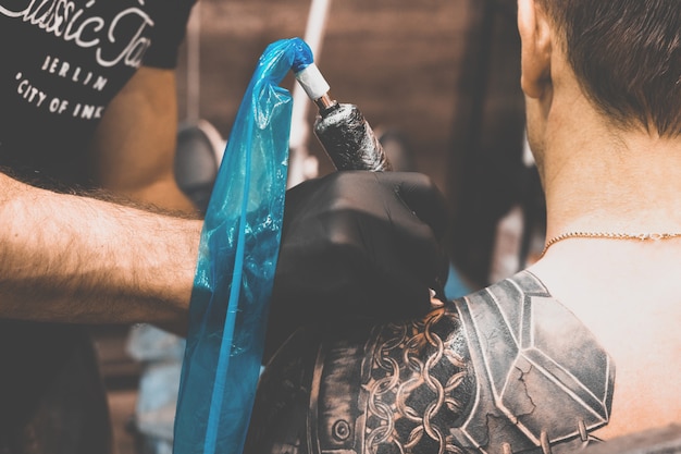 Salone del tatuaggio Il maestro del tatuaggio sta tatuando un uomo sulla sua spalla. Macchinetta per tatuaggi, sicurezza e igiene sul lavoro. Primo piano, colorato, tatuatore