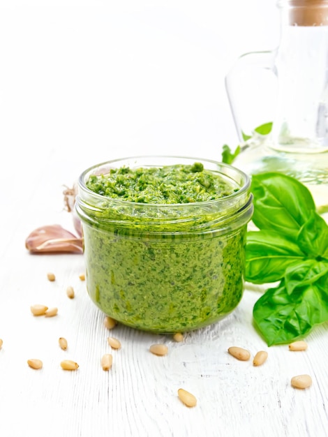 Salsa al pesto in un barattolo di vetro, basilico, pinoli, aglio e olio d'oliva in una caraffa sullo sfondo di una tavola di legno bianca