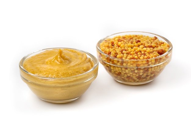 Salsa gialla di senape in una ciotola trasparente e semi di senape sott'aceto isolati su uno sfondo bianco.