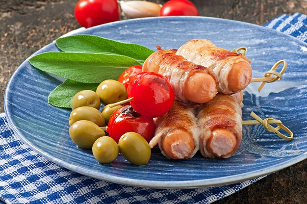 Salsicce alla griglia avvolte in strisce di pancetta con pomodori e foglie di salvia