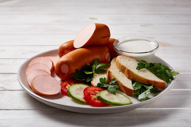 Salsicce con verdure ed erbe aromatiche guarnire con salse e pane su un piatto in ceramica grigia su un tavolo di legno bianco.