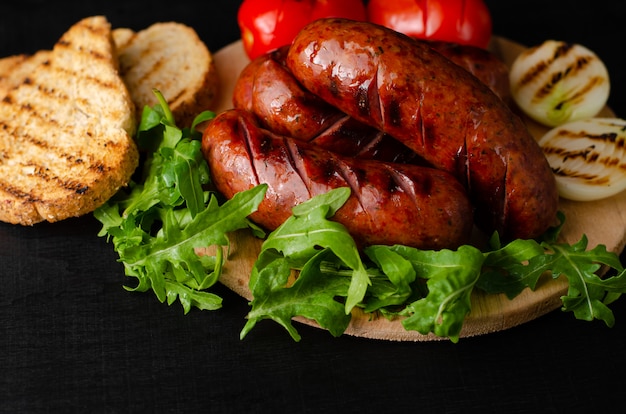 Salsicce di maiale alla griglia con rucola e verdure su sfondo nero. .