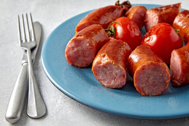 Salsiccia di maiale tagliata a pezzi in un piatto blu con pomodori e posate su uno sfondo di pietra grigio chiaro