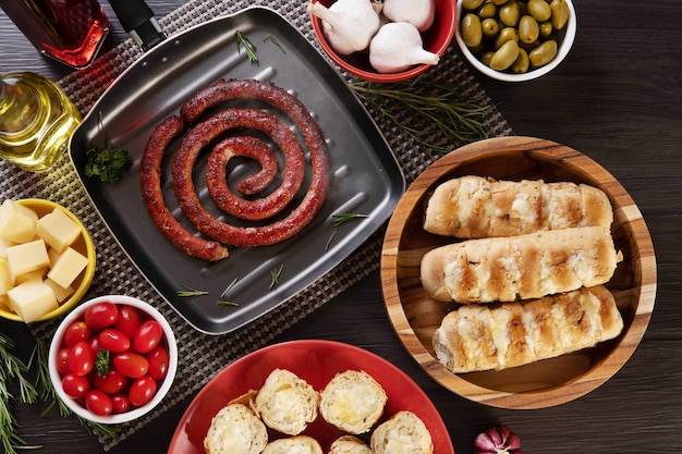 Salsiccia e pane all'aglio su un piatto rosso sul tavolo barbecue con antipasti, formaggio, rosmarino, olive e pomodorini. Vista dall'alto.