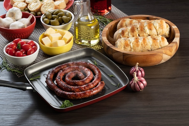 Salsiccia e pane all'aglio su un piatto rosso sul tavolo barbecue con antipasti, formaggio, rosmarino, olive e pomodorini.