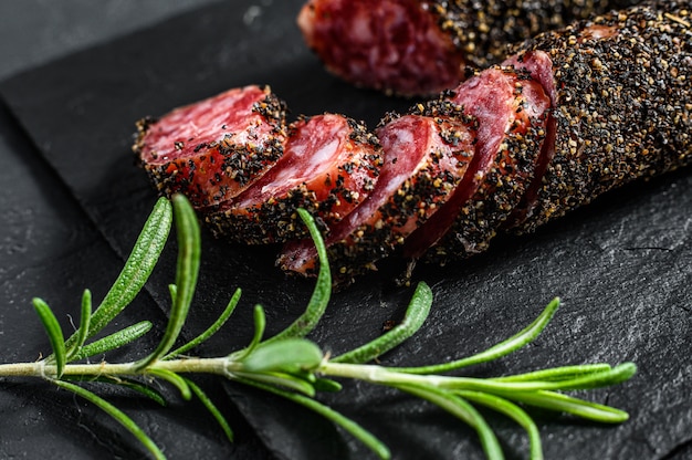 Salsiccia Fuet tagliata a fette su una lastra di ardesia nera con rosmarino. Sfondo nero. Vista dall'alto