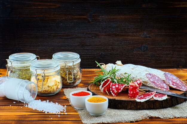 Salsiccia stagionata affettata con spezie e un rametto di rosmarino su fondo rustico in legno scuro.