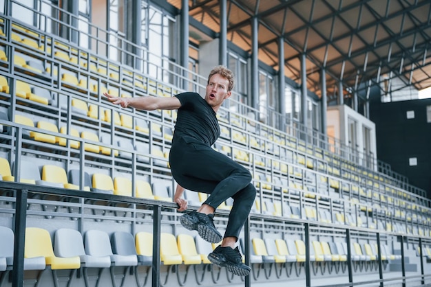 Saltare e fare parkour sulle gradinate Giovane sportivo in camicia nera e pantaloni all'aperto durante il giorno