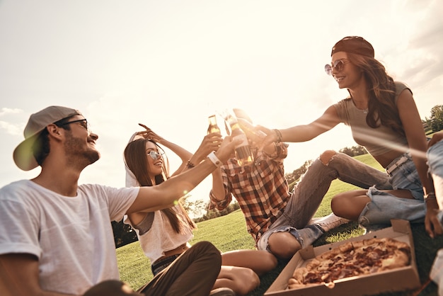 Saluti agli amici! Gruppo di giovani sorridenti in abbigliamento casual che tostano con bottiglie di birra mentre si godono un picnic all'aperto