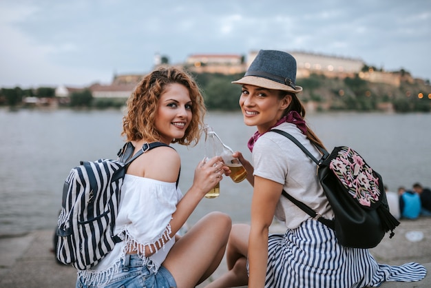 Saluti! Due ragazze alla moda brindano seduti vicino al fiume. Guardando la fotocamera