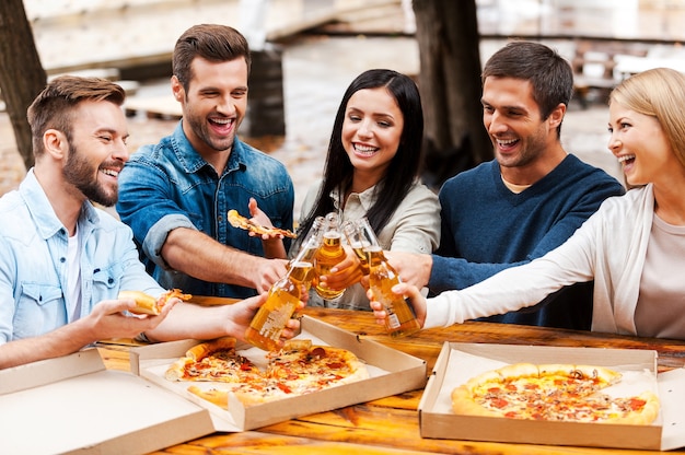 Saluti! Gruppo di giovani allegri che mangiano pizza e fanno il tifo con la birra stando in piedi all'aperto