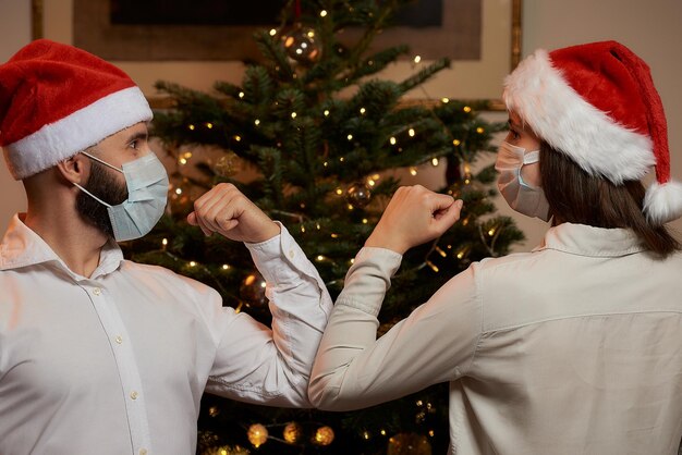 Saluto al gomito per evitare la diffusione del coronavirus COVID19 sullo sfondo di un albero di Natale a casa Uomo e donna felici con maschere mediche e cappelli di Babbo Natale invece di un abbraccio urtano i gomiti