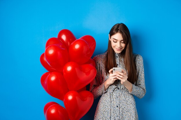 San Valentino. Donna sorridente in abito in piedi vicino a palloncini cuori rossi e guardando smartphone, in piedi su sfondo blu