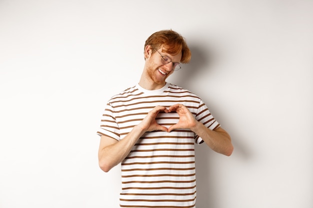 San Valentino. Fidanzato felice con i capelli rossi, sorridente e mostrando il gesto del cuore, ti amo, in piedi su sfondo bianco