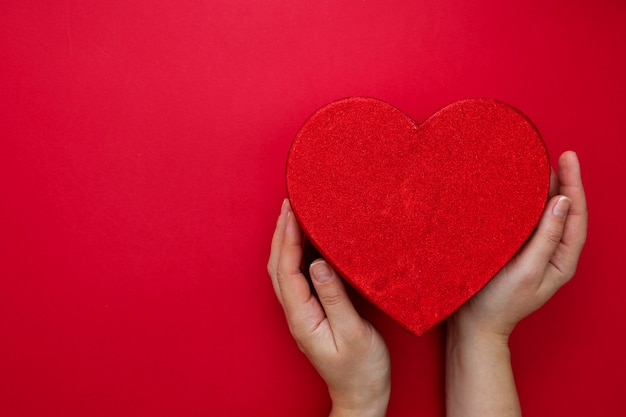 San Valentino mock up. Mani femminili che tengono la scatola del cuore su fondo rosso con lo spazio della copia. Contenitore di regalo rosso astratto.