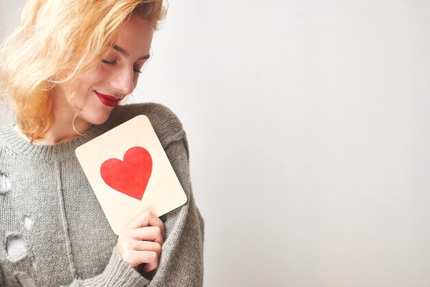 San Valentino. Ragazza in maglione che tiene una carta con un cuore. Su uno sfondo grigio