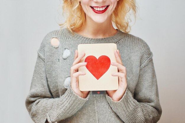 San Valentino. Ragazza in maglione che tiene una carta con un cuore. Su uno sfondo grigio